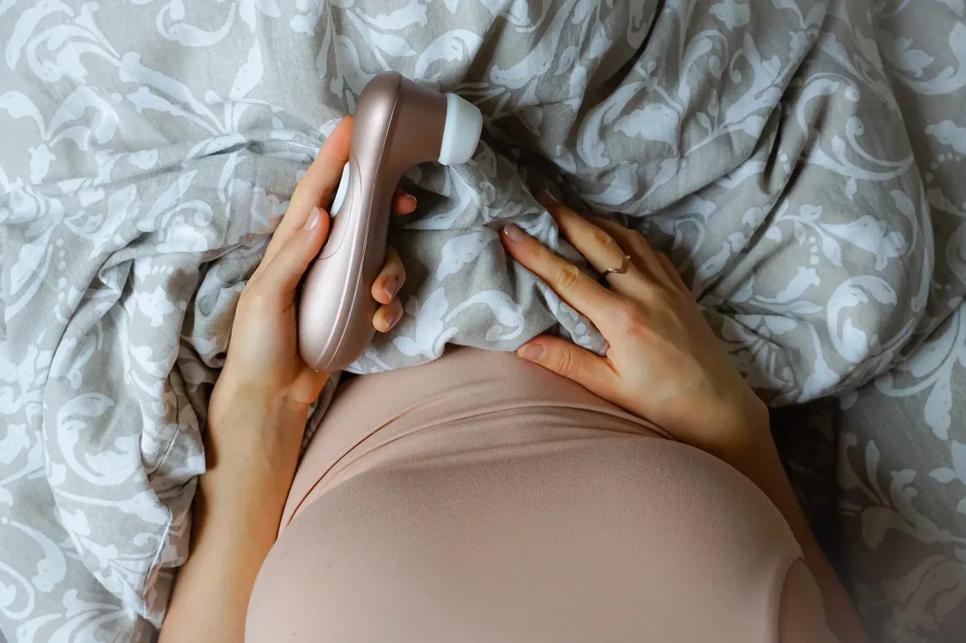 Perspective shot of what a woman would see if sitting in bed and looking down. Woman is in a beige tank top, chest and stomach covered. Lower half is covered by a grey patterned duvet. She is holding a suction toy, it rests on her lap on top of the duvet. 