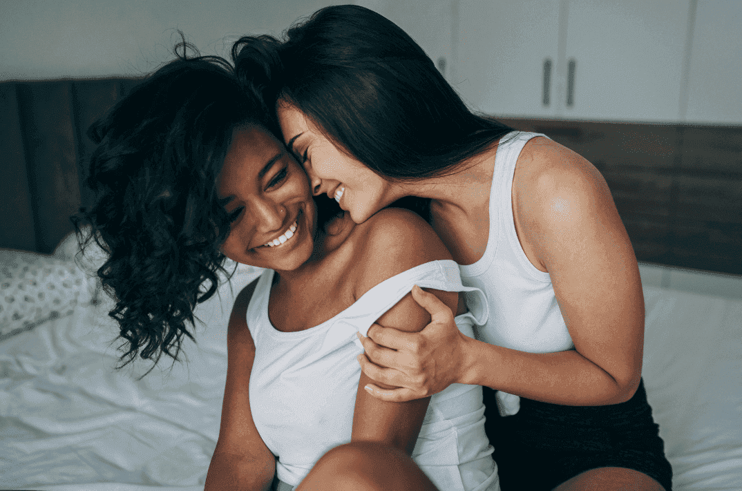 Two women embracing while sitting on the edge of the bed. One woman is brown skinned, smiling, has medium length brown hair, wearing a white tank top. The other woman is fair skinned, cuddles up behind her, forehead pressed against her cheek, smiling. Also in a white tank top. , 