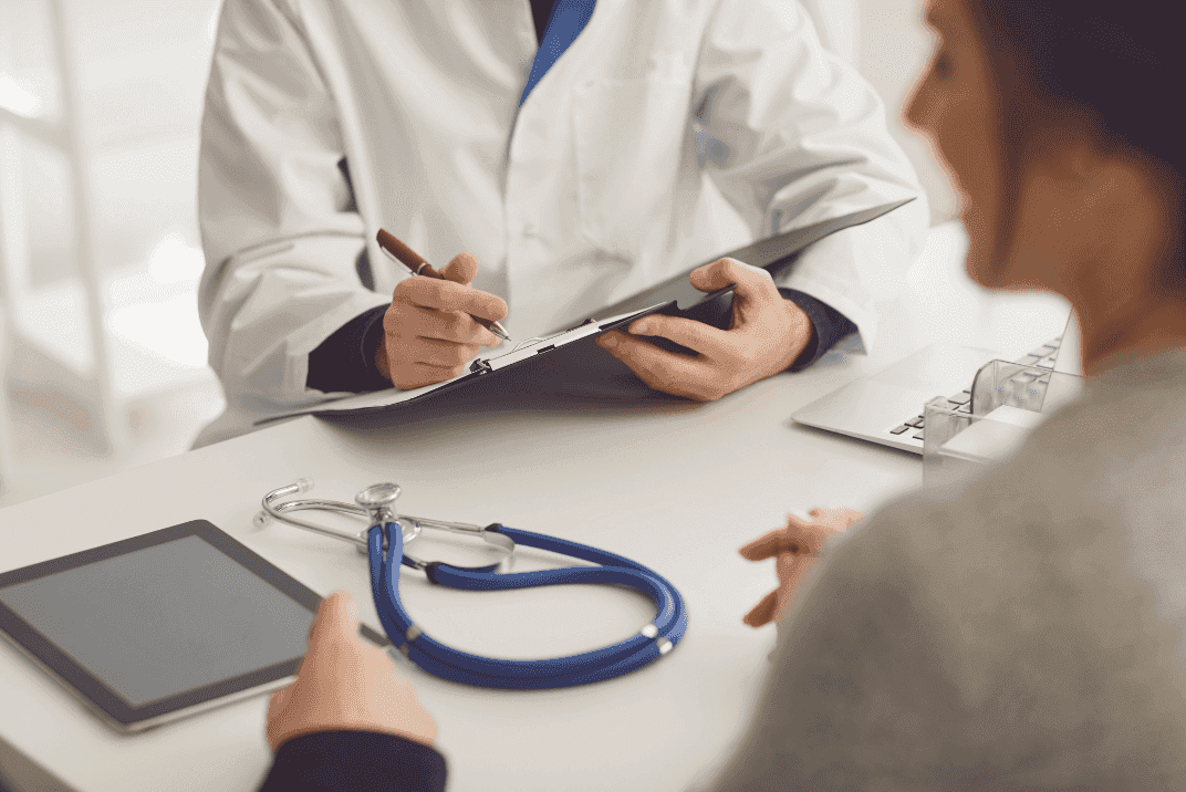 Doctor in a white coat writing on a clipboard, speaking to a woman with her back facing the camera. A man's hand can be seen in the corner, presumably the doctor is counseling a couple. Stethoscope and digital device on the table in front of them. 