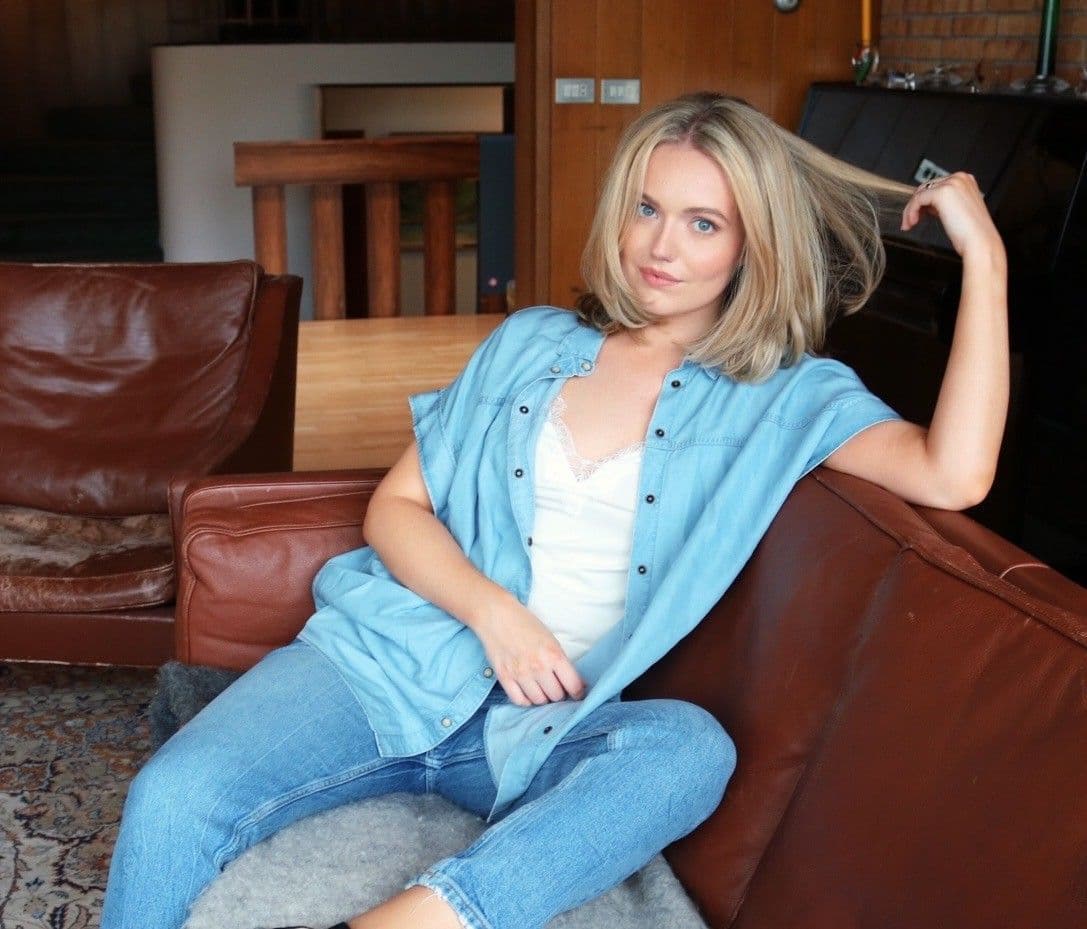 Woman lounging on sofa, strand of her hair in between her fingers, legs up. Wearing an unbuttoned shirt, white undershirt, and jeans. 