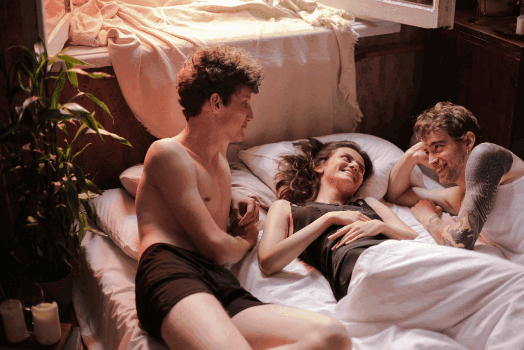 Two men and a woman lounging in bed. Woman is wrapped in a sheet from her lower half down. The group is smiling at each other. One of the men is in his underwear, the other is in a tank top, the photo is cropped to not show his lower half. 