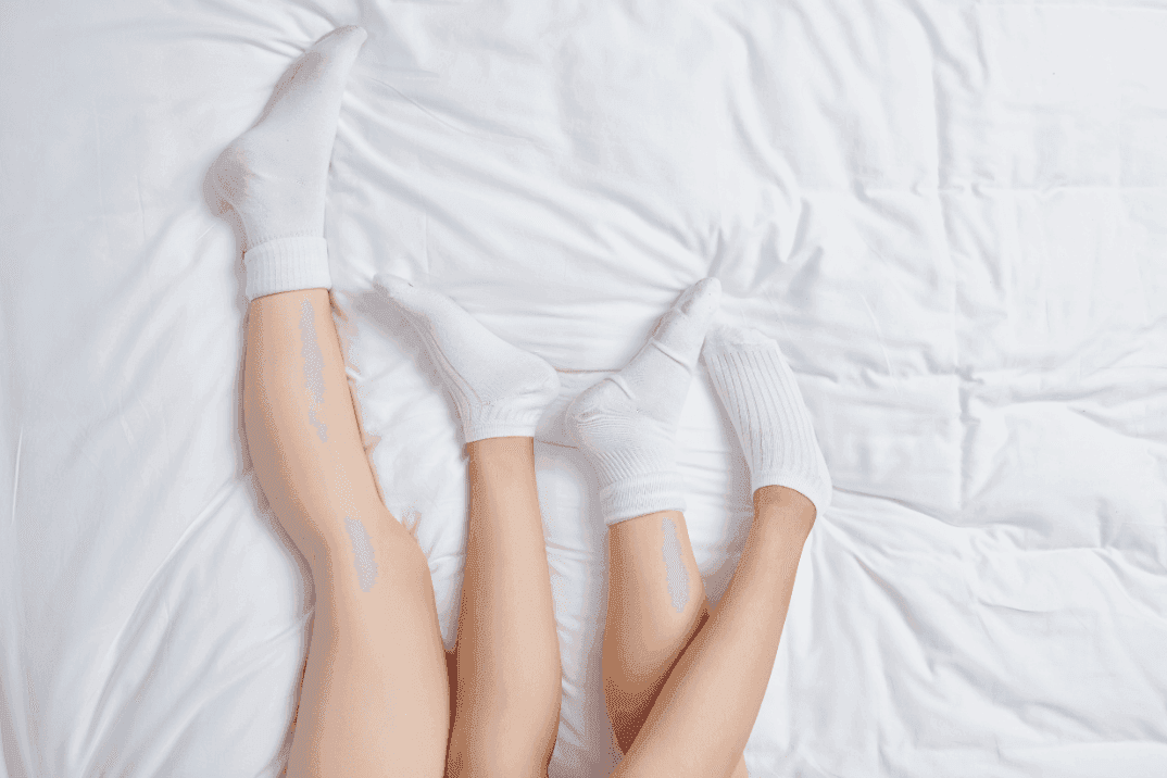 Two women's legs intertwined in bed, both wearing white ankle socks. On top of a white bed sheet. 