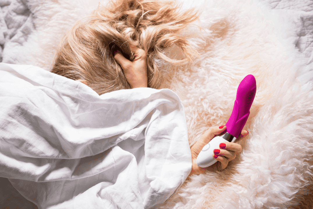 The top of a blonde woman's head is peaking over a sheet, her hand clutches her hair. Her other hand is holding a pink vibrator. She has pink painted nails and is laying on a fuzzy bedspread with a white sheet wrapped around her. 