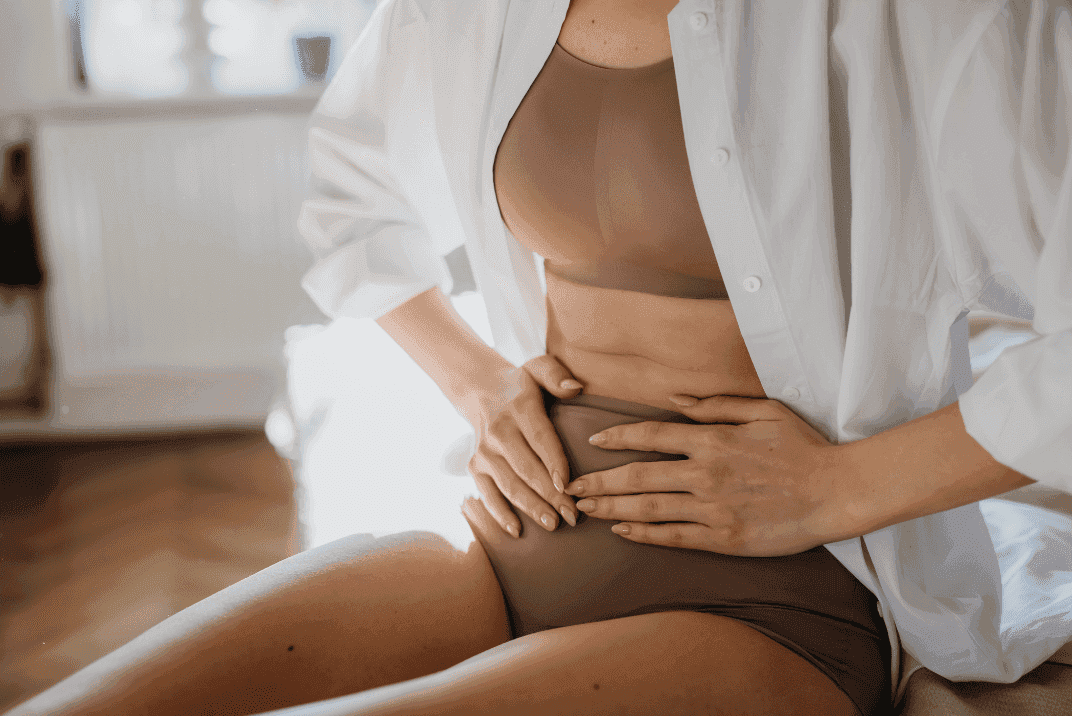 Woman in a two piece underwear set with unbuttoned white shirt. She is sitting up, hands pressed against her stomach. 